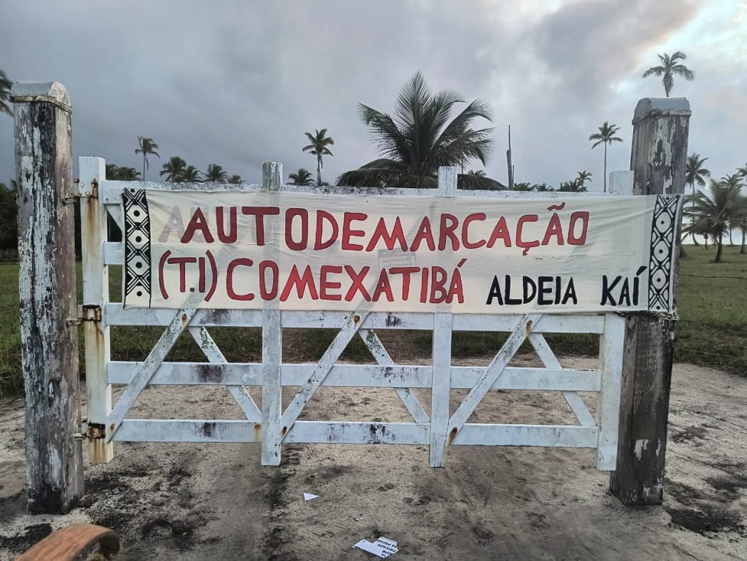 Porteira com faixa onde se lê: Autodemarcação (T.I.) Comexatibá, Aldeia Kaí.